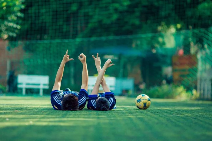 Ảnh bóng đá với hai cầu thủ nằm trên sân cỏ cạnh quả bóng trong không gian ngoài trời xanh mát